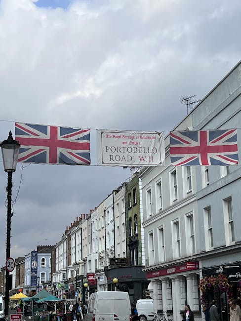A busy street scene in Notting Hill showing a row of white and pastel-colored terraced buildings with shopfronts, some with awnings and signage. The street is filled with pedestrians and parked vehicl