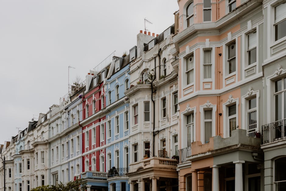 A busy street scene in Notting Hill showing a row of white and pastel-colored terraced buildings with shopfronts, some with awnings and signage. The street is filled with pedestrians and parked vehicl